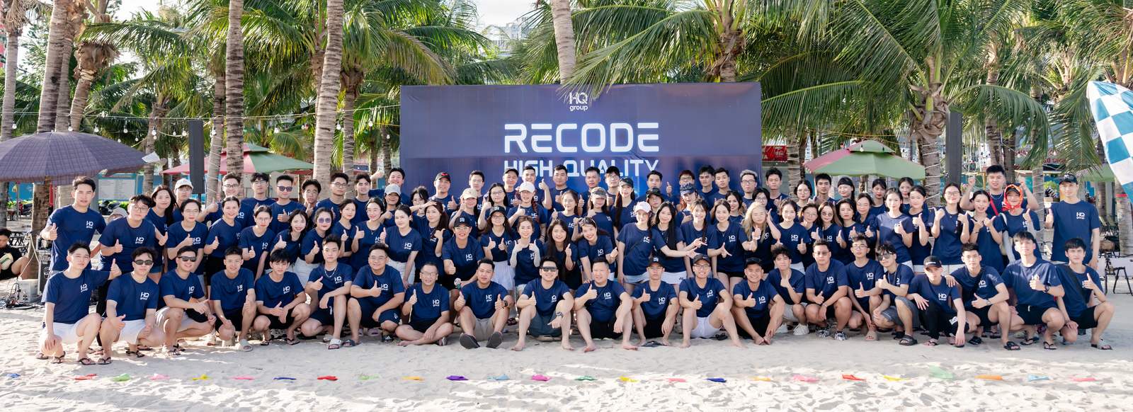 The HQ Group team gathered on a beach in matching navy T-shirts, in front of a Recode High Quality banner.
